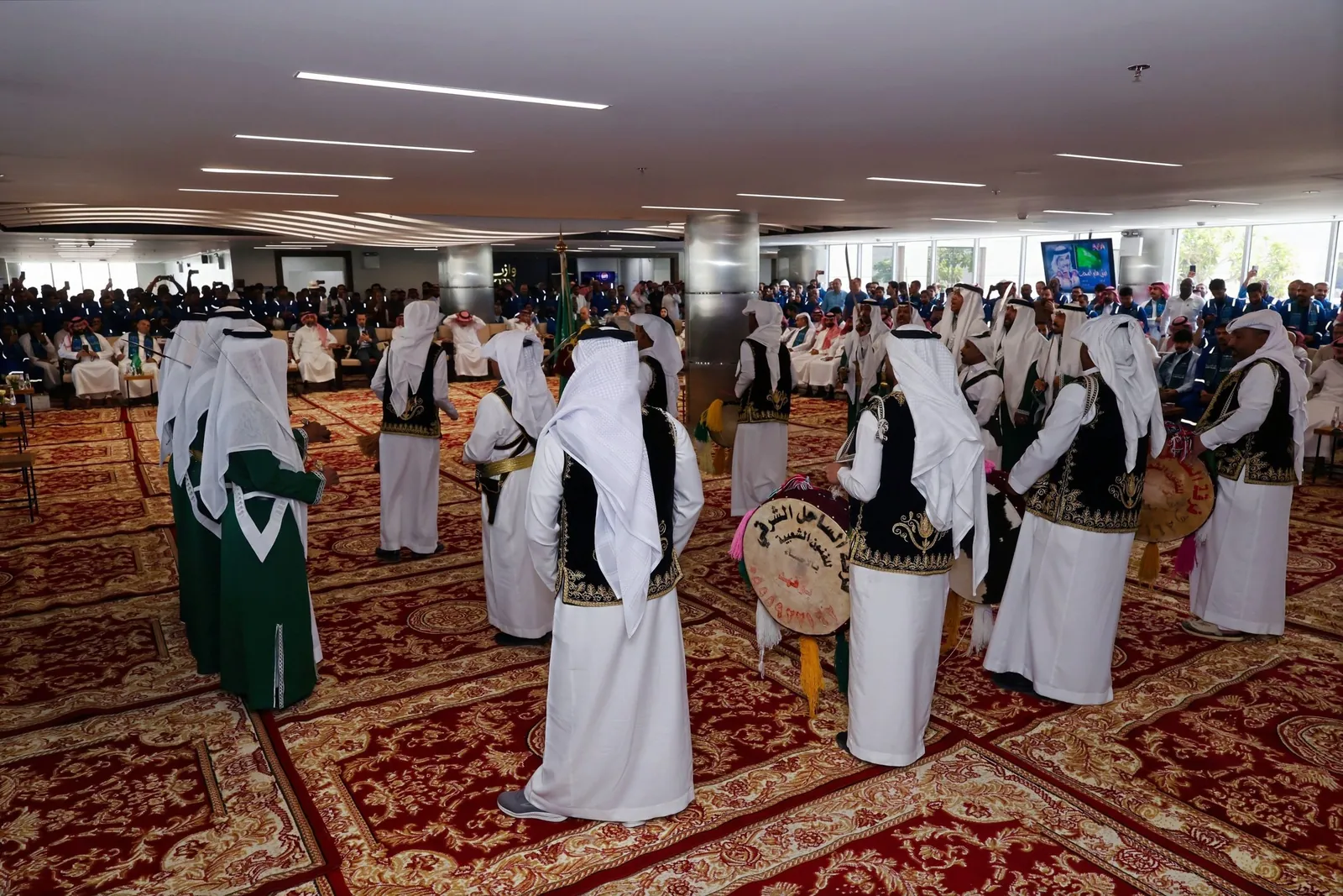 Folk troupe circle with drums on majlis carpets, full crowd watching